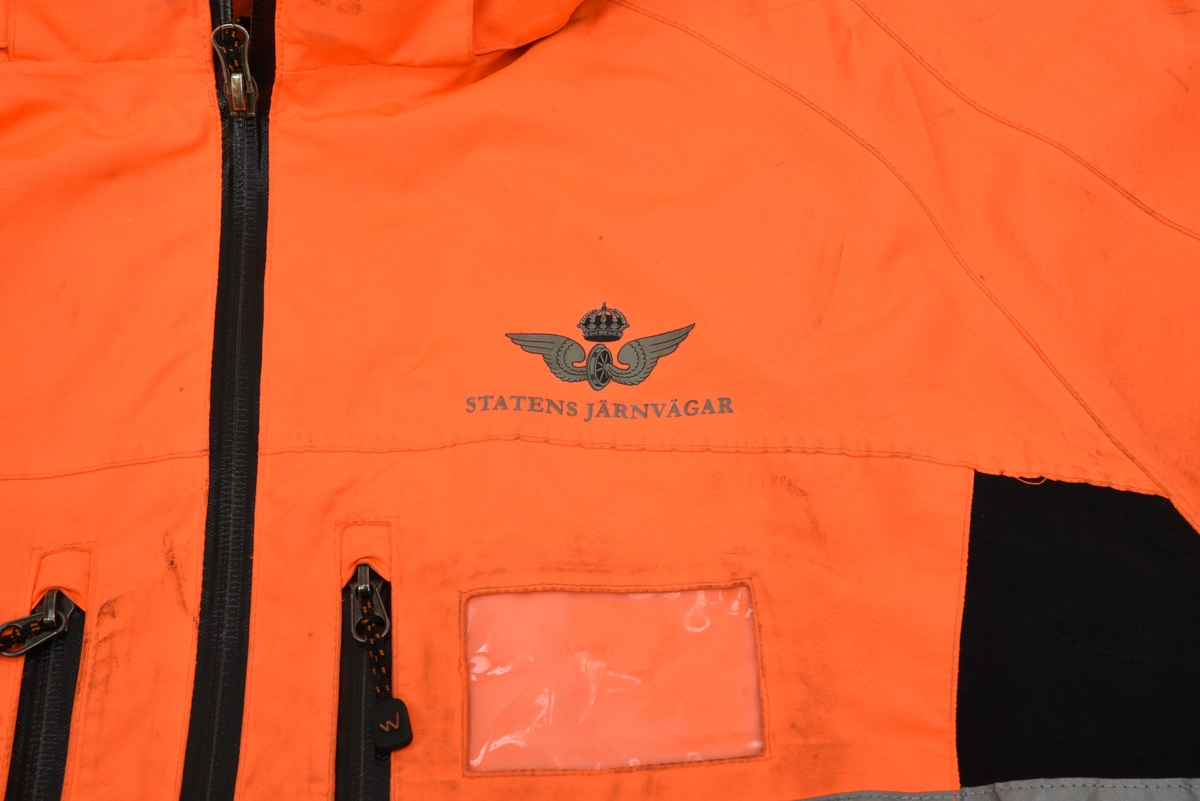 En arbetsjacka, varseljacka, storlek XL. Jackan är orange-aktig i färgen (typiska varselfärgen) och har två  reflexlinjer som går runt hela jackan. På slitytor som armbågar och rumpa har jackan svart textil. Jackan har tre dragkedje försedda fickor på utvändigt.