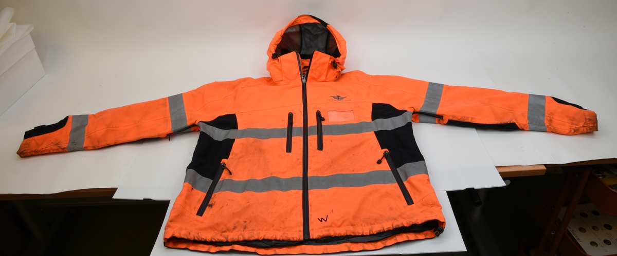 En arbetsjacka, varseljacka, storlek XL. Jackan är orange-aktig i färgen (typiska varselfärgen) och har två  reflexlinjer som går runt hela jackan. På slitytor som armbågar och rumpa har jackan svart textil. Jackan har tre dragkedje försedda fickor på utvändigt.