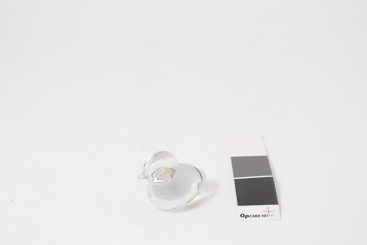 Figur i klart glass, formet som en fugl/and, Merkelapp med produsentlogo på siden.