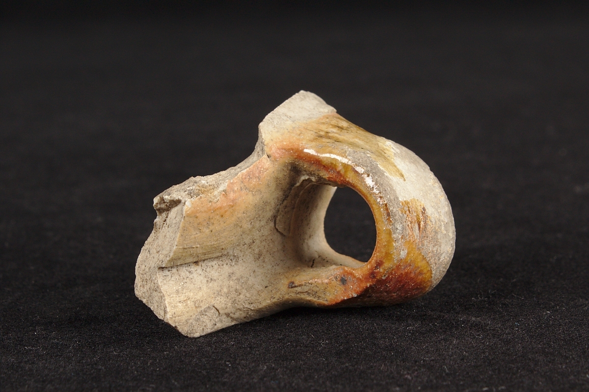 Skärva av drejat och glaserat stengods. Fragment av ett kärl. Överkant med öra.