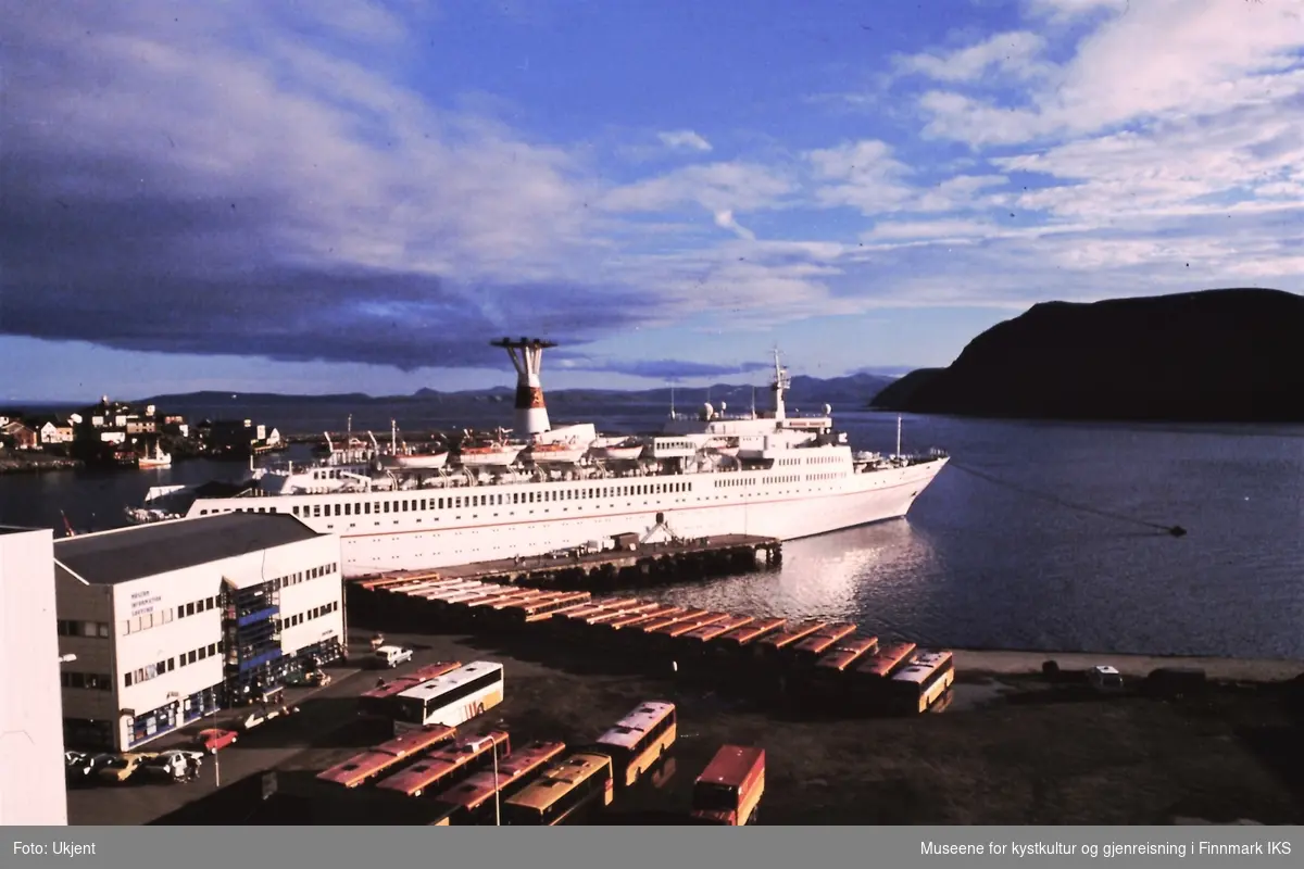 Cruisebåt ''Maxim Gorki' ligger ved kaia i Honningsvåg. Flere busser står parkert. Til venstre Nordkapphuset med museet, turistinformasjon og suvenirbutikk.