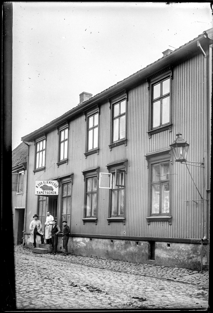 Kongens gate 14 i Moss  ifølge Adressebok for Fredrikshald, Fredrikstad, Moss, Sarpsborg 1911-12.
N. Chr. Kjæmperud Tapetsererforretning. Nils Chr. Kjæmperud f. 1888 i Modum. I folketellinga for 1910 har han bosted i Løkkegata 14a.
En av viserguttene på fotografiet kan være Karl Kjæmperud f. 1894 i Modum.

Siden hadde Josef Bentzen forretning i lokalene (ifølge MBIFs liste).

Kongensgate 14: «På ettermiddagen kom det et stille tog av trauste arbeidere i arbeidsklær, og ofte med matspann, gjennom gata på hjemvei etter endt arbeidsdag på Cellulosen.» Hans Bentzen
«Gata var belagt med firkantet brostein. En gang i uka ble «vår» halvdel feiet med en piasavakost, et ganske tidkrevende arbeid, fordi det gjerne var mye hestemøkk mellom gatesteinene.» Hans Bentzen
«Det fantes verre møkk i nr. 14 en det som hestene la fra seg. Som vanlig på den tida var det også i Bentzegårn en rekke med utedoer, uten elektrisk lys eller oppvarming. Fra tid til annen kom en bonde med hest og vogn for å tømme bingen. Hans Bentzen

«25 ØRE PR. ROTTE. Utedoene og den store søppelkassa av tre plassert nede ved porten, førte til at det ble mye rotter. Vi kunne høre dem når vi satt på do, og vi kunne ofte se dem pile over gårdsplassen» «Min yngre bror fikk ved anledning kjøpt et rottebur. En trapp inne i dette nettingburet førte opp til en fall-lem, der det var plassert noen godbiter. Buret ble plassert i «dogangen», og i løpet av en natt ble buret overbefolket» « I løpet av en uke ble det 50 i alt. Min bror fikk 25 øre av far for hver rotte og var godt fornøyd med «akkorden». Hans Bentzen………………………(Det finnes et rottebur i Halden fra Øvre magasin.)
(Det var minst fem leiligheter i Kongensgate 14) se folketelling 1910