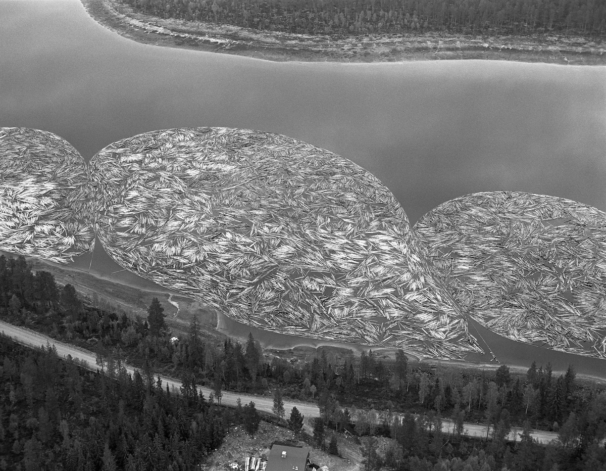 Fortøyde tømmerbommer ved land i Valmen, ei vik i den nordvestre enden av Osensjøen, der sjøen har avløp til elva Søndre Osa. Nordre Osen, Åmot kommune, Hedmark. Fotografiet er tatt fra fly. Veier langs vannet. Hus.