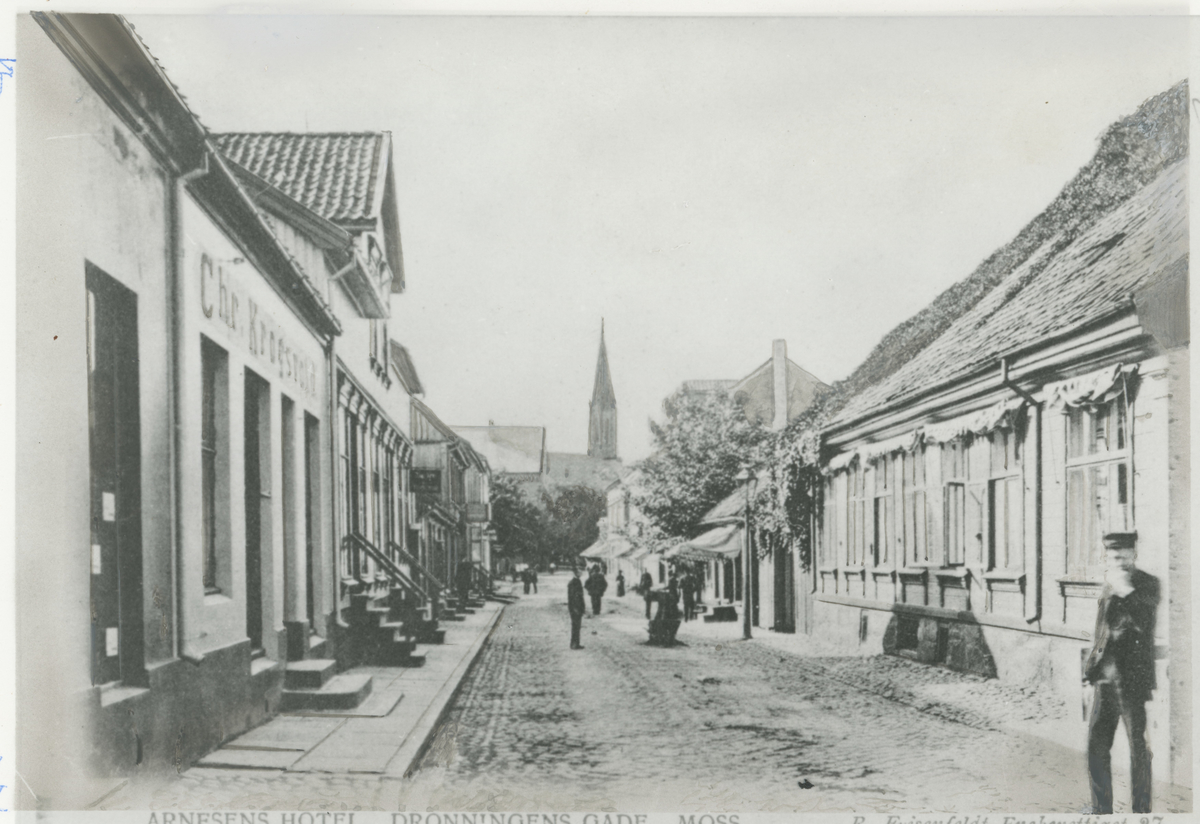 Ca. 1903. Fotograf har stått like utenfor Arnesens hotell i Dronningens gate 3.
Detaljer: Til venstre grosserer Chr. Krogsvolds forretning. Til høyre forsiden av restauranten til Arnesens Hotel.
Historikk: Det meste på venstre side står i dag. Alt på høyre siden er borte.    
Tekst på bildet: “Arnesens Hotel, Dronningens gade, Moss”, “R. Frisenfeldt Eneberettiger 27”