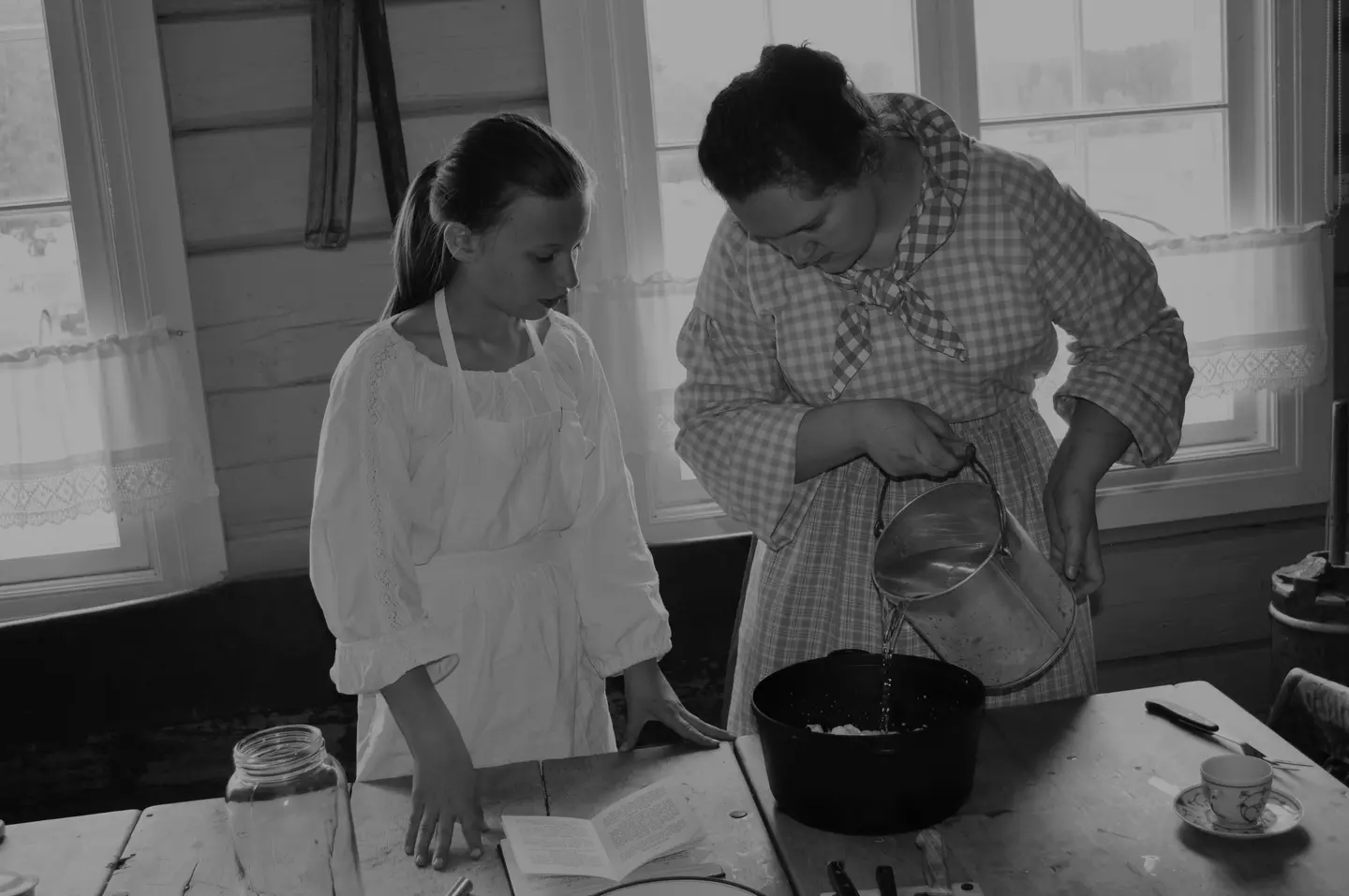 Fotografi i svart-hvitt. Fotografiet viser matlaging på kjøkkenet i Gjersøyen under arrangementet Liv i stuene. En ung kvinne viser en jente hvordan å lage en matrett. Kvinnen har på rutete forkle, rutete skjorte og rutete tørkle. Jenta har på hvit bluse og hvitt forkle. De er fokuserte på matlagingen.