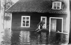 Översvämning.
Ett bostadshus står till viss del under vatten.
Två män utanför, varav den ene bär den andre på ryggen.