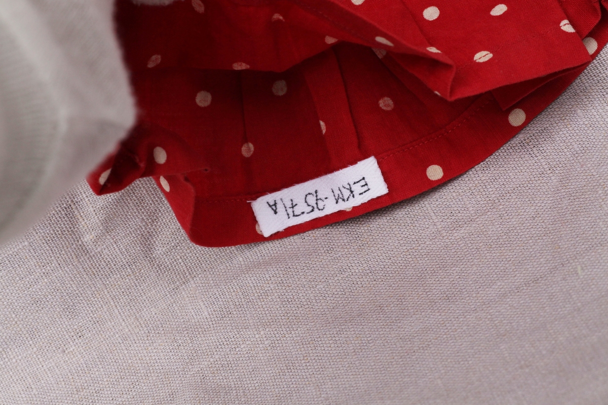 Tre enkle dukkekjoler, to rødprikkete (a og b) og en blårutet (c), en stråhatt (d), et bånd (e). Rød kjole (b) uten ermer, med hvite kanter.