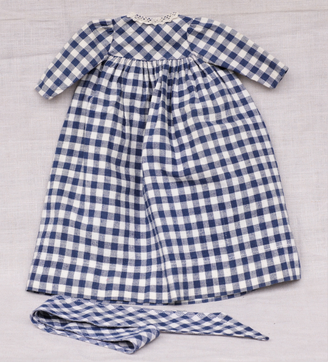 Tre enkle dukkekjoler, to rødprikkete (a og b) og en blårutet (c), en stråhatt (d), et bånd (e). Rød kjole (b) uten ermer, med hvite kanter.