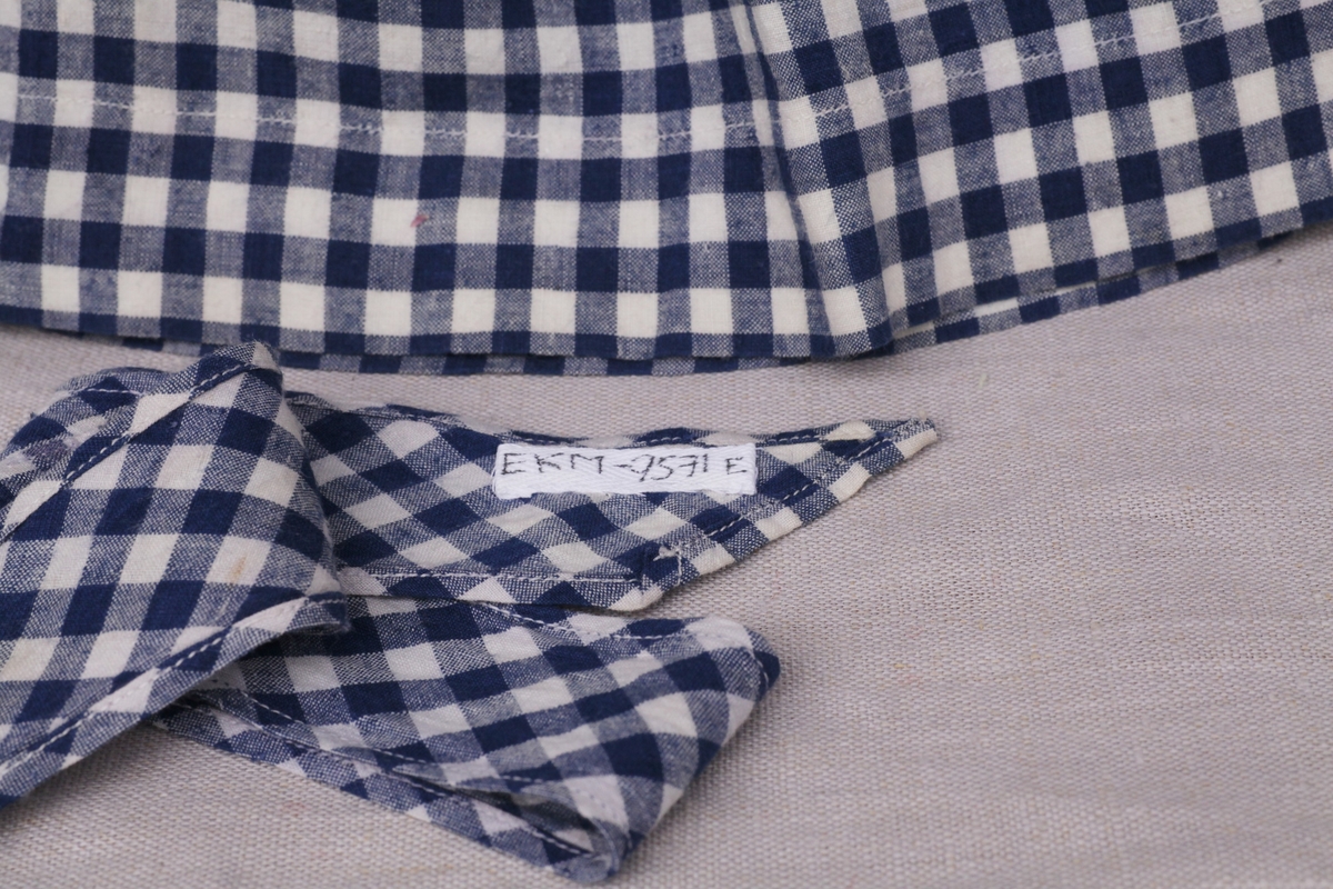 Tre enkle dukkekjoler, to rødprikkete (a og b) og en blårutet (c), en stråhatt (d), et bånd (e). Rød kjole (b) uten ermer, med hvite kanter.