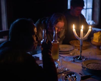 Julemiddag i Vestaberg under Jul i stuene. Vestaberg er husmannsplassen på Odalstunet. Et trearmet lys på bordet. I forgrunn ser vi en mann som nyter julemiddagen. Ved bordet sitter en ung jente og en ung gutt.