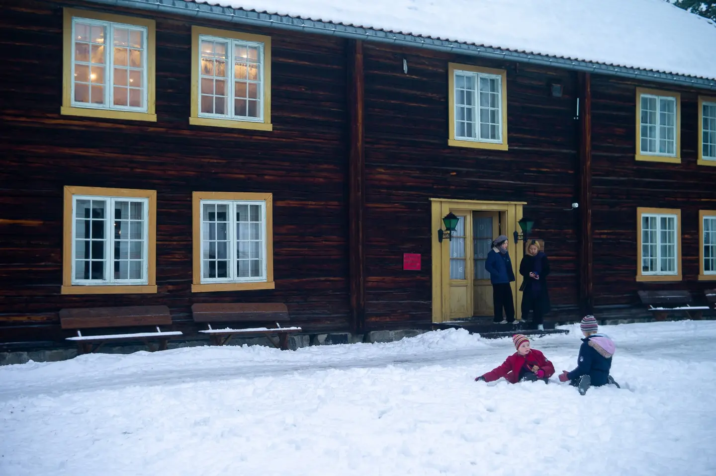 Fotografi tatt av Gjersøyen-bygget under Jul i stuene. Et staselig tømmerhus med store fine vinduer. Vinduskarmer og dør er sennepsgul. To etasjer. Utenfor bygget er det to voksne mennesker som har en samtale ved inngangsdøren, og to barn som leker i snøen.