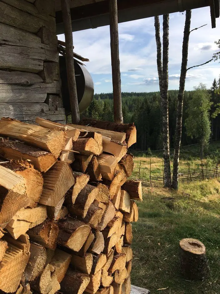 Bildet av en vedstabel på Skålbergsætra. Veden er stablet opp inntil hovedbygget på seteren. Bildet er tatt slik at det viser hjørnet av bygget. I bakgrunnen ser man setervangen i sola.