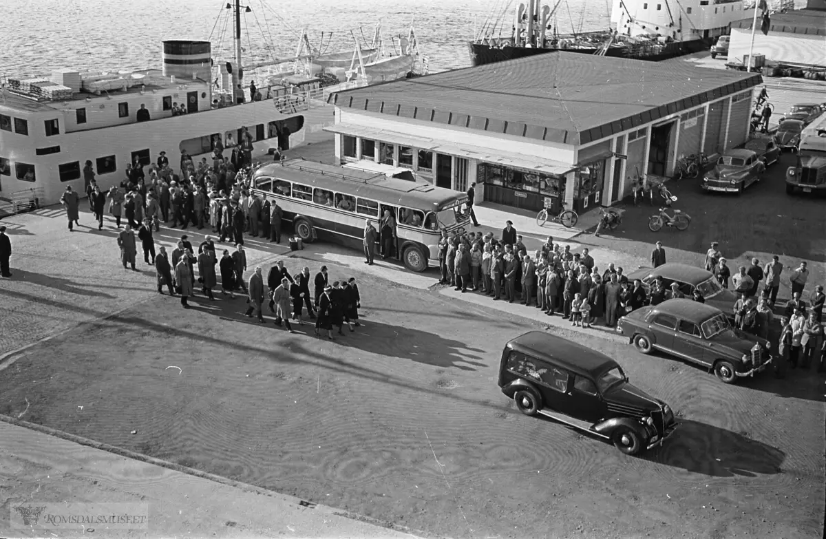 "September 1958"."Oksviks gravferd".Fra begravelsen til Olav Oksvik f.07.05.1887 d.16.09.1958..Båten som ligger til kai er M/S Romsdal, som gikk i rute Åndalsnes-Molde-Ålesund i tidsrommet 1950-1962. Den ble solgt i 1962..Bussen på bildet er sannsynligvis T-8685 Scania-Vabis, 1953-modell, med karosseri fra Bussbygg, Hovdenakken. Bussen tilhørte Aarø Automobilselskap (Auto) og den var lakkert i gult og mørk blå. Aarø Auto hadde ruter Molde-Kristiansund over Gjemnes, og ruta startet og avsluttet ved Storkaia i Molde, der det var korrespondanse med båter til Åndalsnes og Ålesund. .Begravelsesbilen er en Ford V8 1936 med registreringsnummer T-2167...På høyre side av bygningen en Dodge eller Chrysler personbil 1946-48-modell og til høyre for den utsnitt av en Scania-Vabis semitrailer.