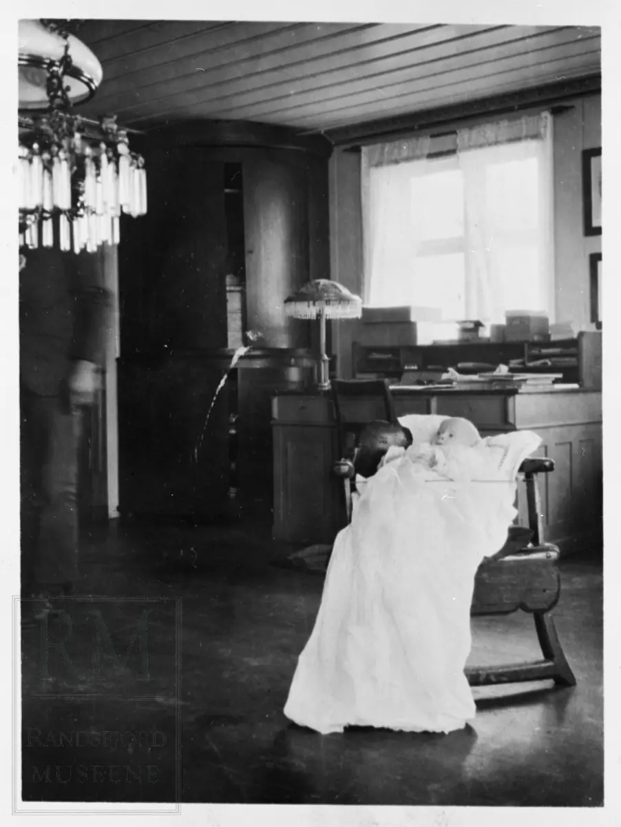 Babyen Marius Gisleberg i dåpskjole, plassert i en stol. En skrivepult ved et vindu i bakgrunnen.