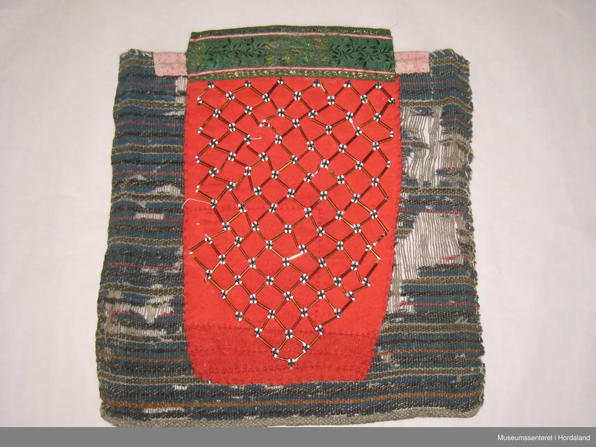 Stor brystduk med perlenett sydd på raudt ullstoff. Nede er det sydd på tre remser av eit anna raudt ullstoff. Montert på eit vove tøystykke der innslagstrådane av ull er borte fleire stader. Renning av heimespunne lin. Kasta over på to sider og falda på to sider med hand. Eit rosa stoff er sydd på oppe til forsterking. Grønt silkeband med rosekvarde og gullfarga metalltråder vove inni er montert utanpå det rosa stoffet øvst.