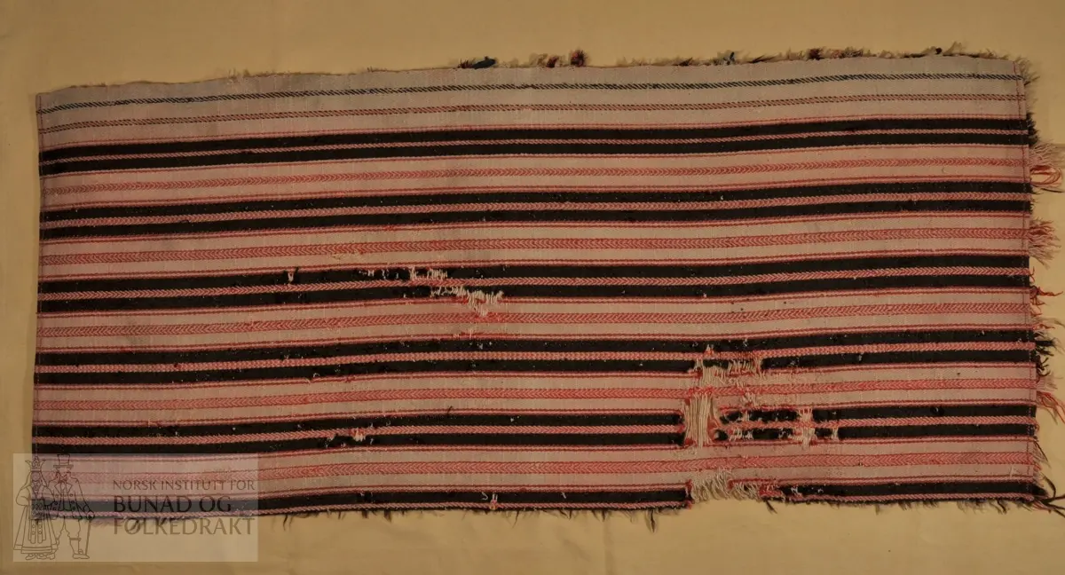 Båtrye, vevd av ull i kypert. Knyttet på floss underveis i veven. Farger i stripene /renninga : Sauesvart, rødrosa og ubleika . 	Innslaga er ubleika ull. Floss i rosa og ubleika ull.