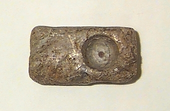 På ena bredsidan streckfigurer (bl.a. ett ansikte med en krona), på den motsatta sidan en hemisfärisk urgröpning. Även en oval urgröpning på ena smalsidan. I huvudliggaren upptagen som gjutform för rektangulärt spänne.