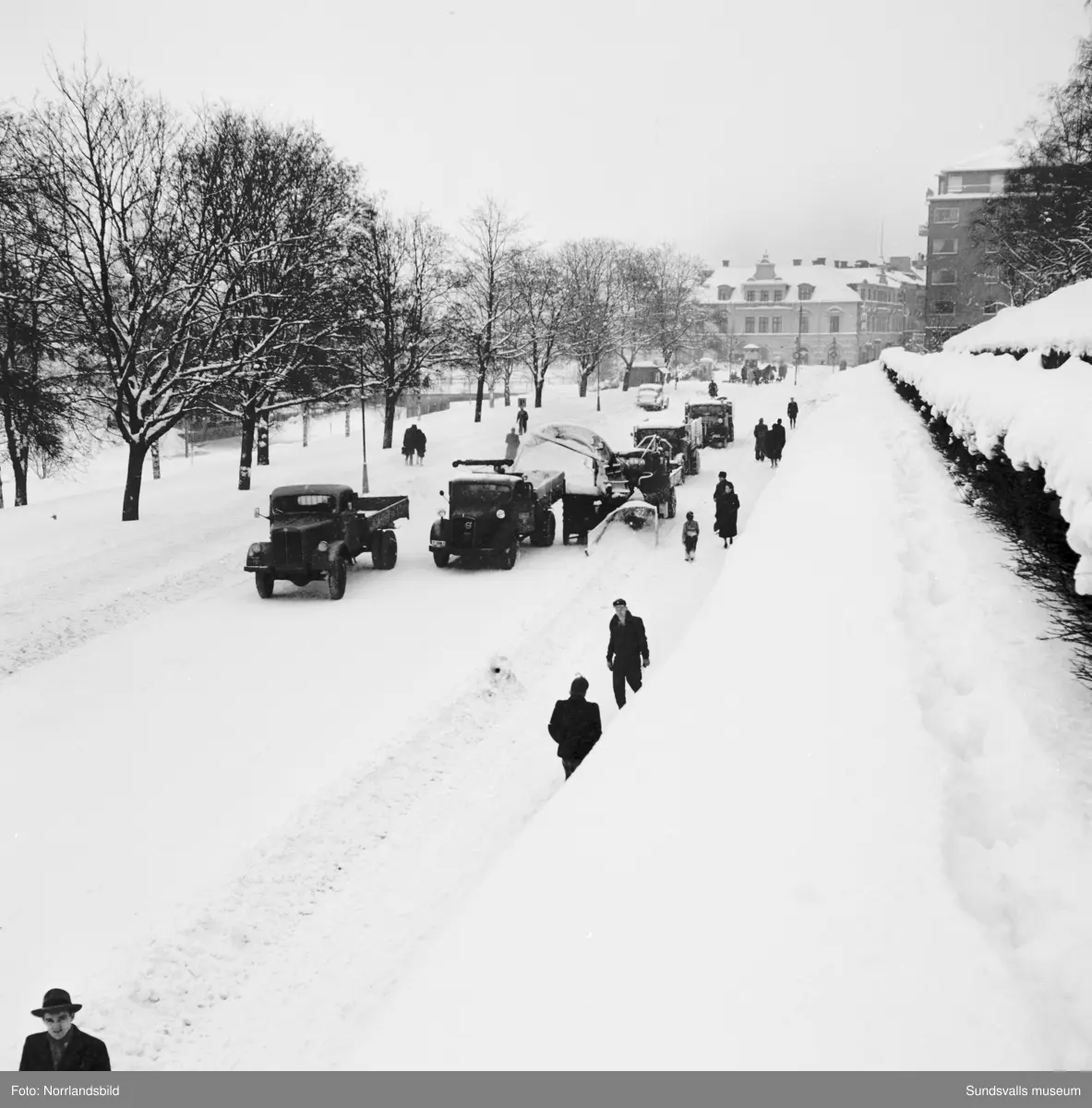 Snöröjning med plog, lastbilar och snöslunga på västra Storgatan. Några barn leker en farlig lek framför snöslungan.