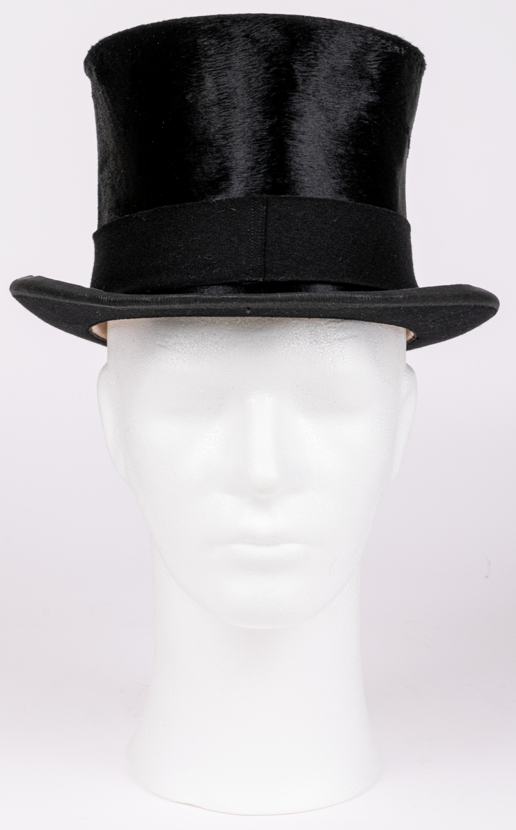 Stormhatt märkt Aktiebolaget J.W Haglund, N. Kungsgatan 1, Gefle. Svart.
Höjd 14cm, brättbredd 4,5cm.
Hattask av svart papp med guldtext på locket. English manufacture of hats. Best London deposed.