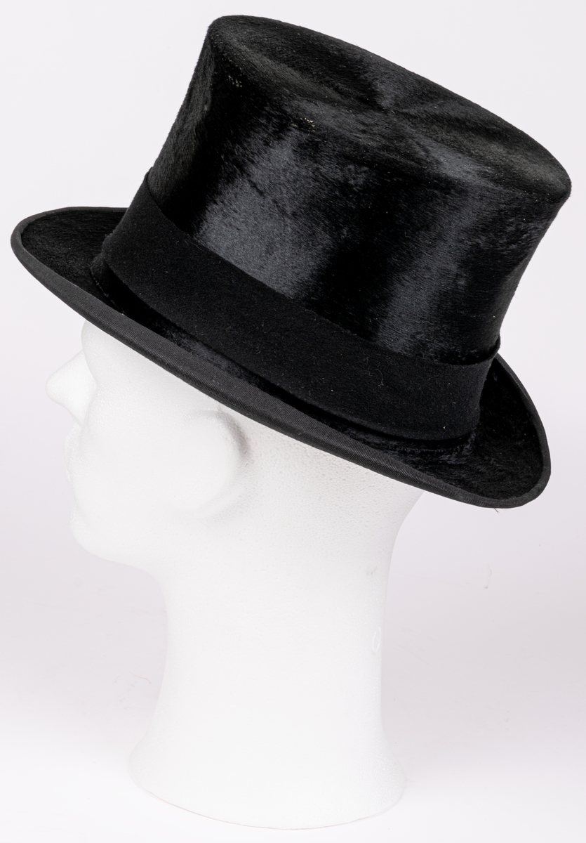 Stormhatt märkt Aktiebolaget J.W Haglund, N. Kungsgatan 1, Gefle. Svart.
Höjd 14cm, brättbredd 4,5cm.
Hattask av svart papp med guldtext på locket. English manufacture of hats. Best London deposed.