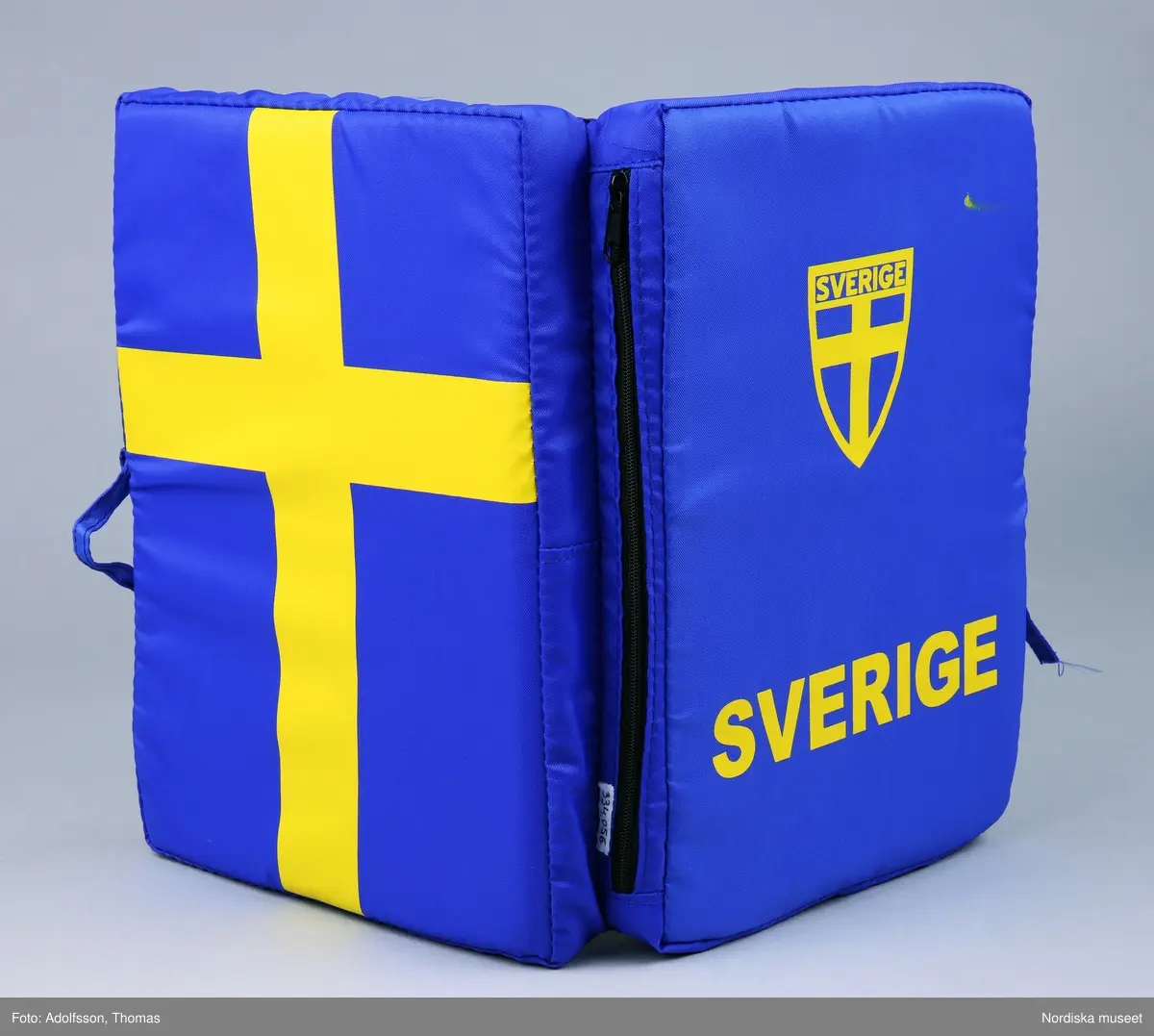 Sittdyna med svenska flaggan som dekor.  Av blått syntetmaterial med fyllning av skumgummi. På framsidan står SVERIGE i gult tryck, därovanför en version av Sveriges landslagsmärke för fotboll med texten Sverige På baksidan en svensk flagga i tryck. Dynan är uppfällbar, själva sittdynan har också svenska flaggan som dekor. Dynan kan knytas till i hopfällt skick. Oanvänd.

/ C Hammarlund-Larsson 2019-01-25