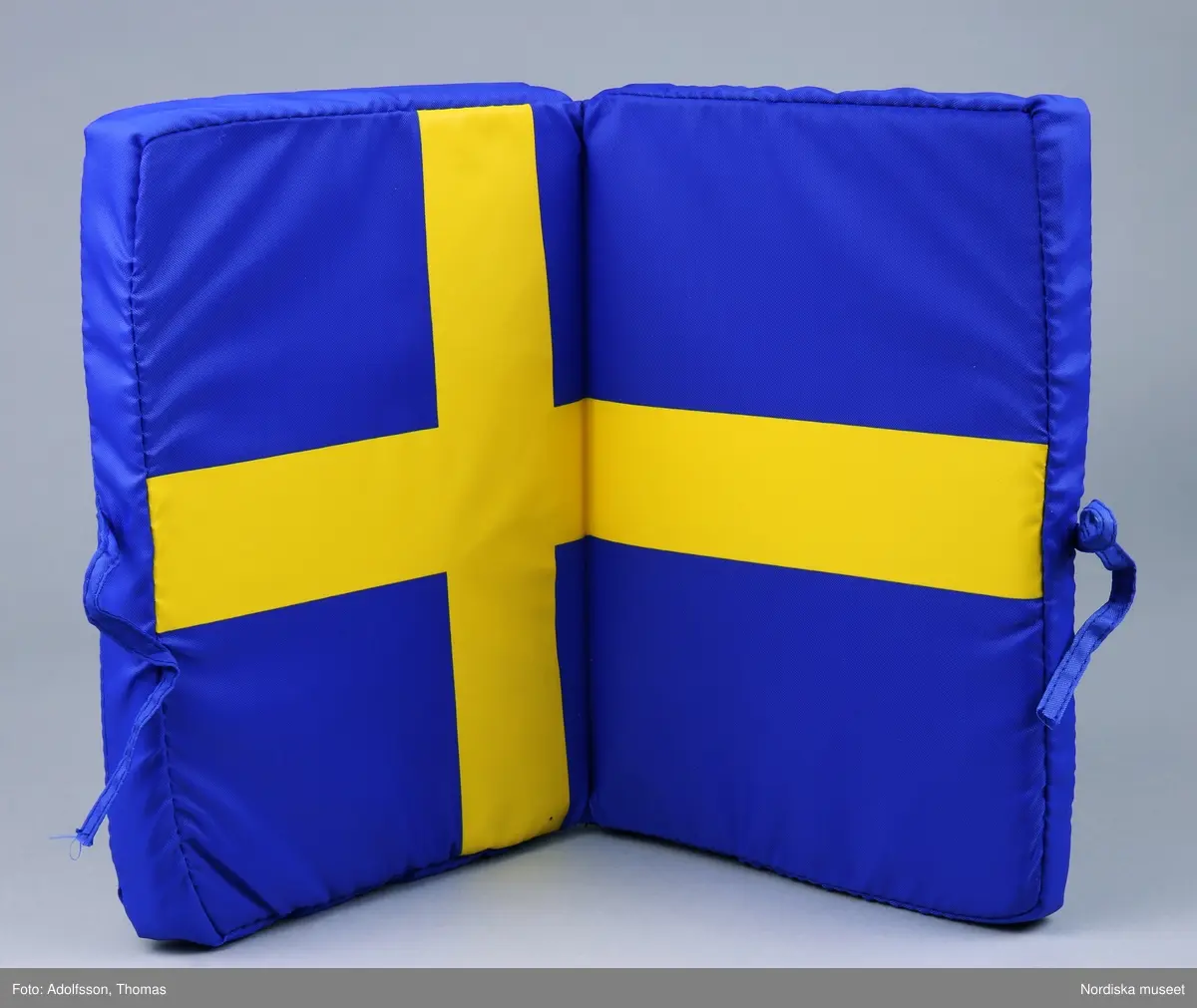 Sittdyna med svenska flaggan som dekor.  Av blått syntetmaterial med fyllning av skumgummi. På framsidan står SVERIGE i gult tryck, därovanför en version av Sveriges landslagsmärke för fotboll med texten Sverige På baksidan en svensk flagga i tryck. Dynan är uppfällbar, själva sittdynan har också svenska flaggan som dekor. Dynan kan knytas till i hopfällt skick. Oanvänd.

/ C Hammarlund-Larsson 2019-01-25