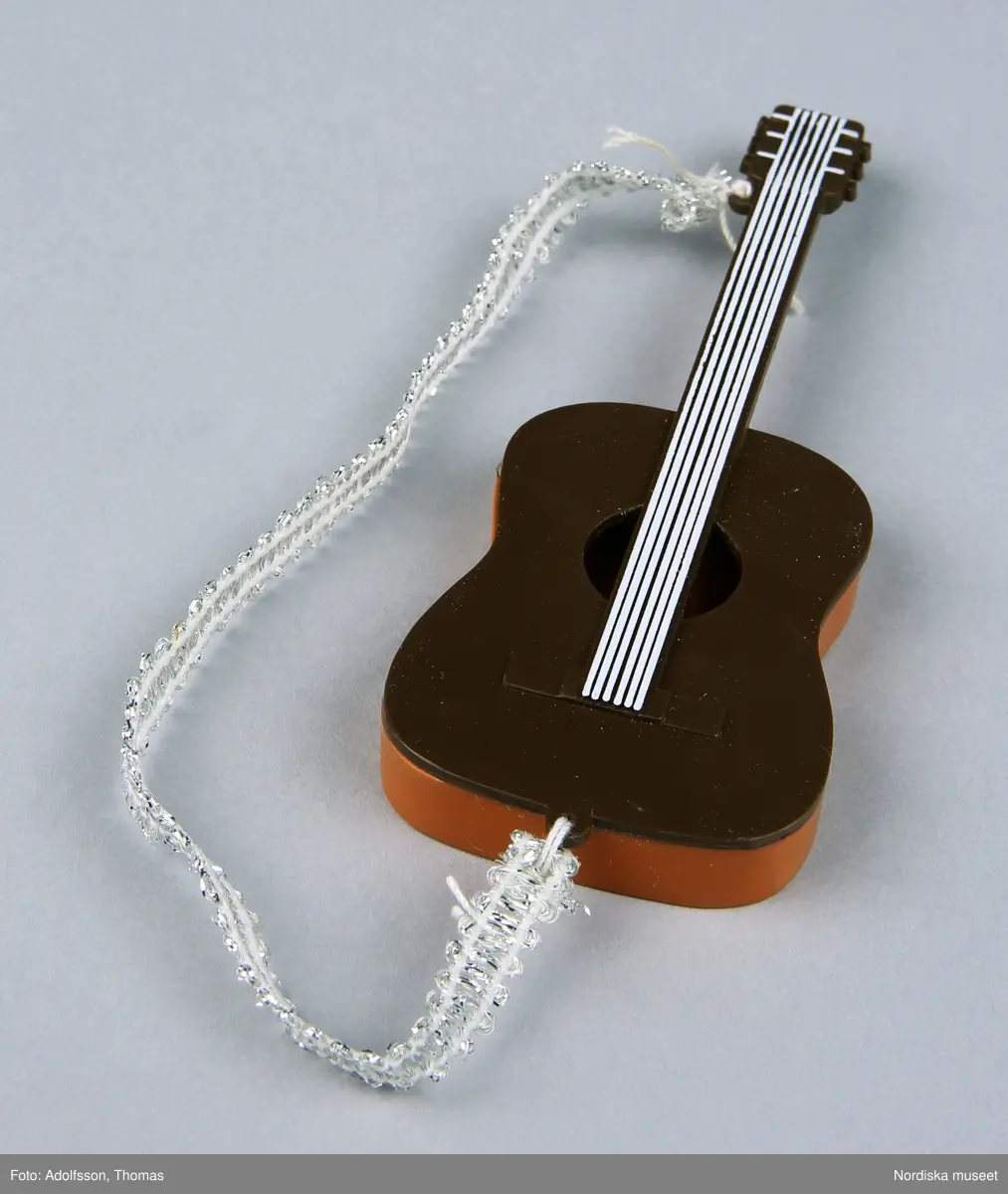 a-b) Två likadana dockgitarrer av plast med ett silverfärgat band av syntet. Den ena hör till dockan Benny, NM0334232 a-e. och den andra till Björn, NM0334233 a-d.