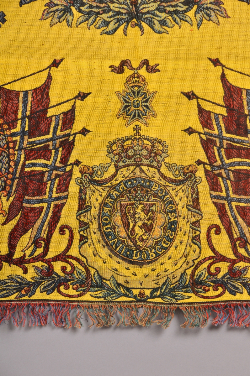 Tekstil i bomull med litt metalltråd. Fleire fargar. Portrett av HaakonVII på midten. I kvart hjørne er der portrett av Kong Haakon, Dronning Maud og Kronprins Olav. Den norkse løva med teksten "Haakon VII Norges Konge", Flagg, ranker og løver m.m. Tafs kring alle kantane. Tafset er ein forlengelse av varp og verft. Tydeleg rett og vrangside. God stand, men noko bleika på den eine sida.