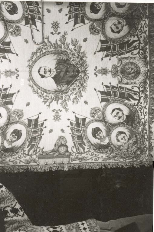 Tekstil i bomull med litt metalltråd. Fleire fargar. Portrett av HaakonVII på midten. I kvart hjørne er der portrett av Kong Haakon, Dronning Maud og Kronprins Olav. Den norkse løva med teksten "Haakon VII Norges Konge", Flagg, ranker og løver m.m. Tafs kring alle kantane. Tafset er ein forlengelse av varp og verft. Tydeleg rett og vrangside. God stand, men noko bleika på den eine sida.