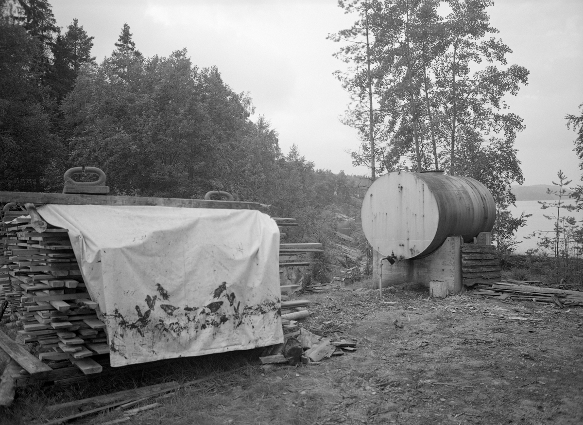 Bordstabel ved Sleppetangen i Spydeberg i Østfold.  Borddstabelen er delvis dekt av en lys presenning.  Til høyre ses en oljetank av stål på fundamenter av betong.  I bakgrunnen skimtes vannflata på Øyeren.  Sleppetangen var oppankringssted for tømmerslep fra Fetsund lenser i nordenden av innsjøen Øyeren.  Her skulle slepene løses opp og tømmeret slippes løst mot nedre Glomma. 