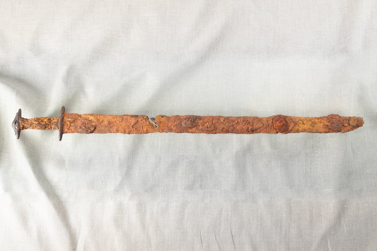 Depåfynd av svärd, cirka 85 cm lång från svärdsknapp till spets. Cirka 20 cm från hjaltet är klingan bruten, ett större brott, och här saknas även en del material från klingan och skidan. Cirka 20 cm av bladets övre del är även det avbrutet, vilket upptäcktes vid flytt från brädan till vattenkar. Handtaget i trä med spår av läder är välbevarat. Svärdsbaljan är i stort sett komplett och den inre stommen av trä syns. Baljan är omslagen med vad som tolkas vara läder och eventuellt kan fragment av textil ses i baljans spets.