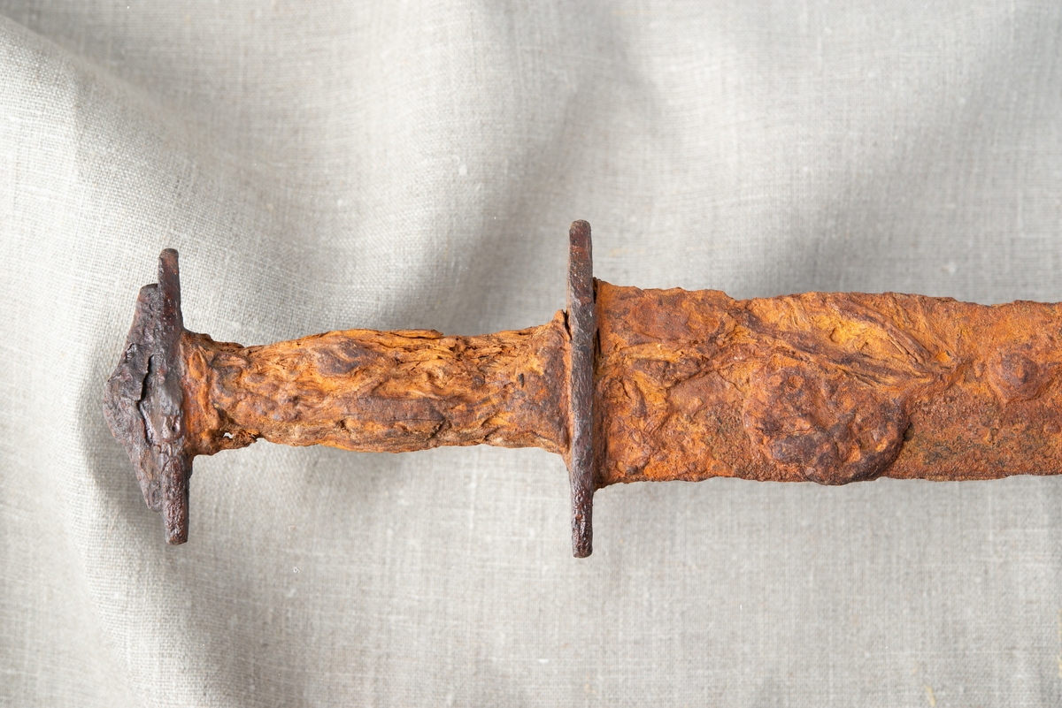 Depåfynd av svärd, cirka 85 cm lång från svärdsknapp till spets. Cirka 20 cm från hjaltet är klingan bruten, ett större brott, och här saknas även en del material från klingan och skidan. Cirka 20 cm av bladets övre del är även det avbrutet, vilket upptäcktes vid flytt från brädan till vattenkar. Handtaget i trä med spår av läder är välbevarat. Svärdsbaljan är i stort sett komplett och den inre stommen av trä syns. Baljan är omslagen med vad som tolkas vara läder och eventuellt kan fragment av textil ses i baljans spets.