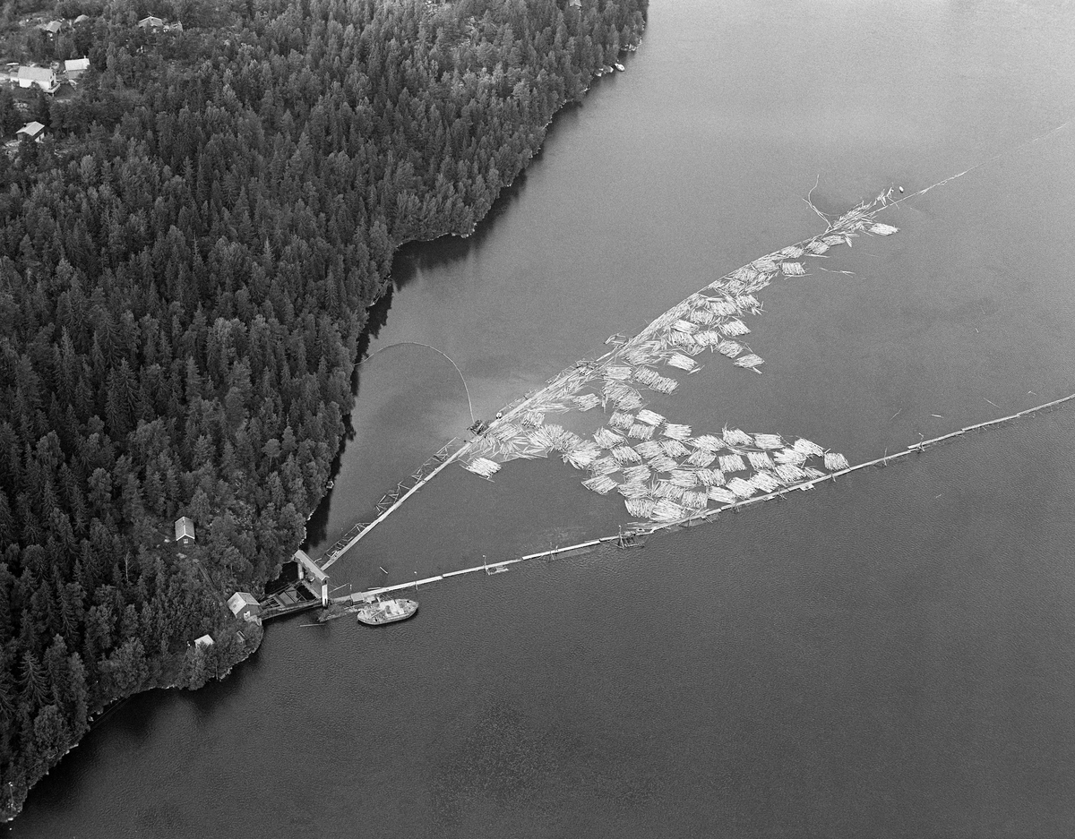 Flyfotografi av lensa som ledet tømmer inn mot tømmerrenna ved Solbergfoss kraftstasjon i grenseområdet mellom Spydeberg og Askim i Østfold.  Fotografiet er tatt fra et punkt omtrent rett over dammen, ned mot tunnelåpningen ved vestre elvebredd.  Vi ser den V-formete lensa, der den nedre delen er ei kraftig stivlense.  Her ligger det en god del «moser» - tømmerbunter.  Vanligvis kom tømmeret løst ned til dette stedet fra Sleppetangen ved Glommas utløp fra innsjøen Øyeren litt høyere oppe i vassdraget.  I perioder da vindretningen var slik at en kunne risikere at løstømmer drev tilbake i innsjøen, hendte det imidlertid, som her, at mosene ble sendt hele.  I slike tilfeller måtte «grimene» (vaierbindslene) tas av før tømmeret ble sendt løst inn i tømmertunnelen.  Ved inngangen til tunnelen ser vi en slags betongportal, en justerbar terskel som gjorde det mulig å regulere vannføringa og strømhastigheten i tømmertunnelen med varierende vannstand i hovedvassdraget.  Fotografiet er tatt i 1985, det siste året det foregikk kommersiell tømmerfløting i dette vassdraget.