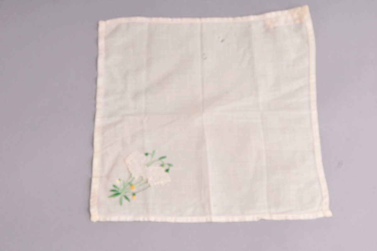 Kvadratiske lommetørkle i tynnt bommullstøy. Blomsterbroderi og applikerte blomar nede i eine hjørnet.Smal holfald kring kanten. Lommetørkledet er brodert med maskin.