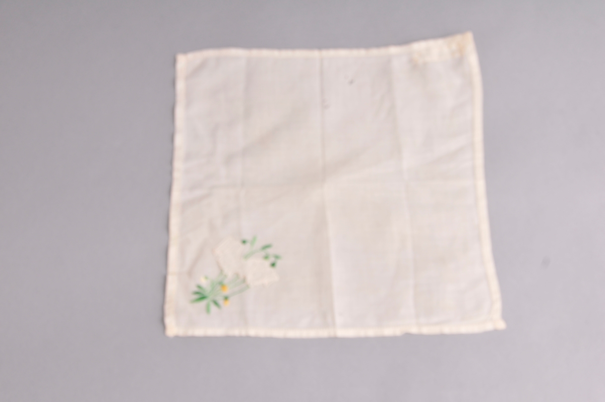 Kvadratiske lommetørkle i tynnt bommullstøy. Blomsterbroderi og applikerte blomar nede i eine hjørnet.Smal holfald kring kanten. Lommetørkledet er brodert med maskin.
