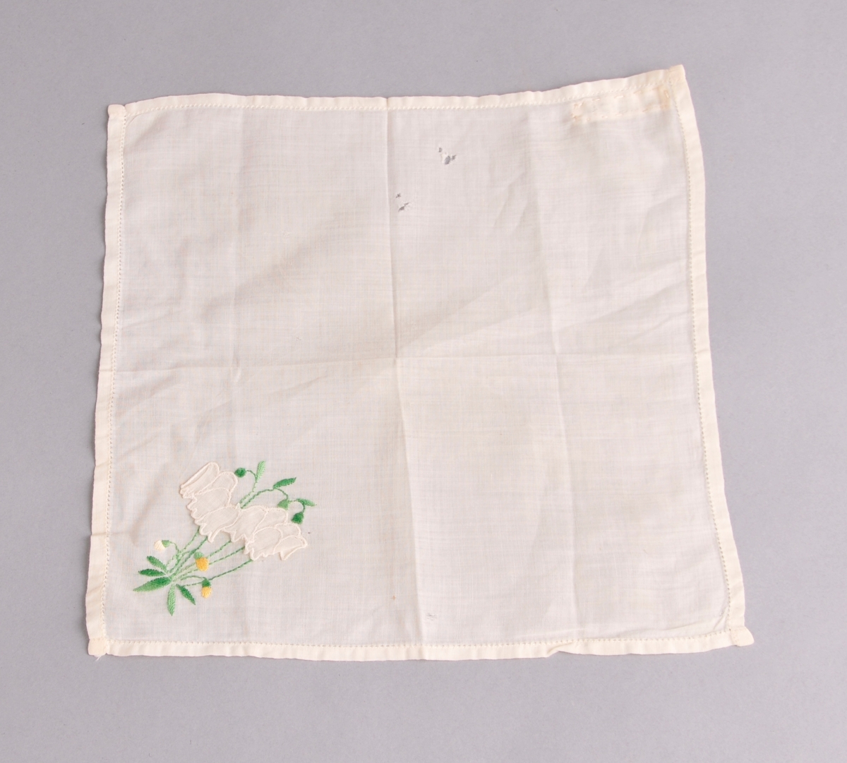 Kvadratiske lommetørkle i tynnt bommullstøy. Blomsterbroderi og applikerte blomar nede i eine hjørnet.Smal holfald kring kanten. Lommetørkledet er brodert med maskin.
