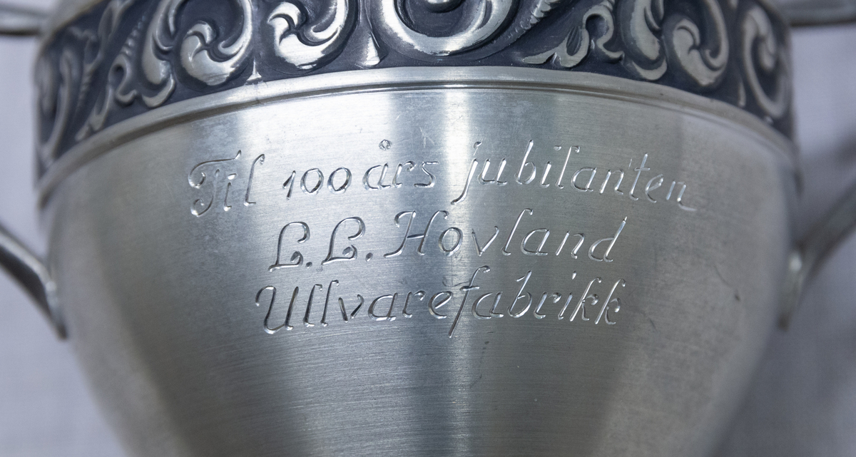 Pokalforma vase med to sidehankar. Dekorfelt rundt vasen med utstansa (?) rosemalingsmønster.