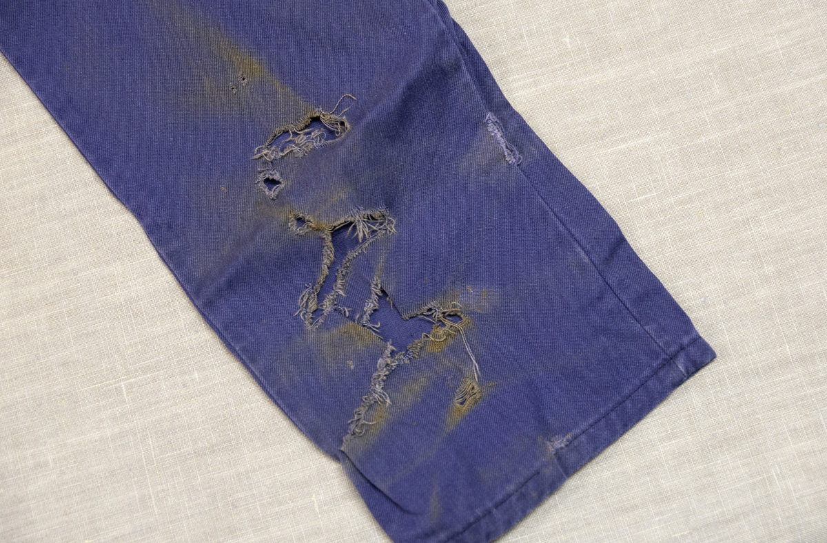 Todelt dress i mørk blå bomullsdenim. Storleik 48 (båe delene). Glidelås i metall framme i jakke, knepping med metallknappar i buksegylf. To brystlommer på jakka, to stikklommer i buksa. Innside av bukselinning i tynt nylonstoff (?). Buksa har produsentmerket laminert fast i høgre lomme og jakka har tilsvarande på innsida bak høgre brystlomme. Vaskeanvisning 60 gr. 

Buksa har fleire større og mindre hol, særleg nede på venstre buksebein, - både framme og bak. Om desse er forårsaka av varme eller syre er ikkje kartlagt, men fargen tyder på høg varme. Det er ikkje gjort forsøk på reparasjon av hola. Stoffet er ikkje flammehemma.