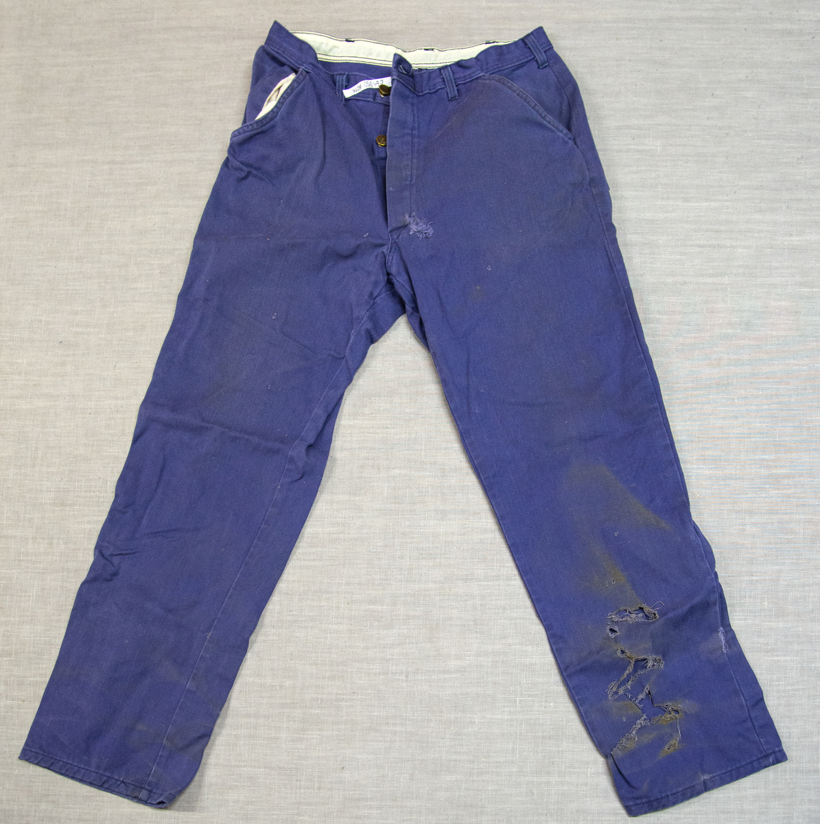 Todelt dress i mørk blå bomullsdenim. Storleik 48 (båe delene). Glidelås i metall framme i jakke, knepping med metallknappar i buksegylf. To brystlommer på jakka, to stikklommer i buksa. Innside av bukselinning i tynt nylonstoff (?). Buksa har produsentmerket laminert fast i høgre lomme og jakka har tilsvarande på innsida bak høgre brystlomme. Vaskeanvisning 60 gr. 

Buksa har fleire større og mindre hol, særleg nede på venstre buksebein, - både framme og bak. Om desse er forårsaka av varme eller syre er ikkje kartlagt, men fargen tyder på høg varme. Det er ikkje gjort forsøk på reparasjon av hola. Stoffet er ikkje flammehemma.