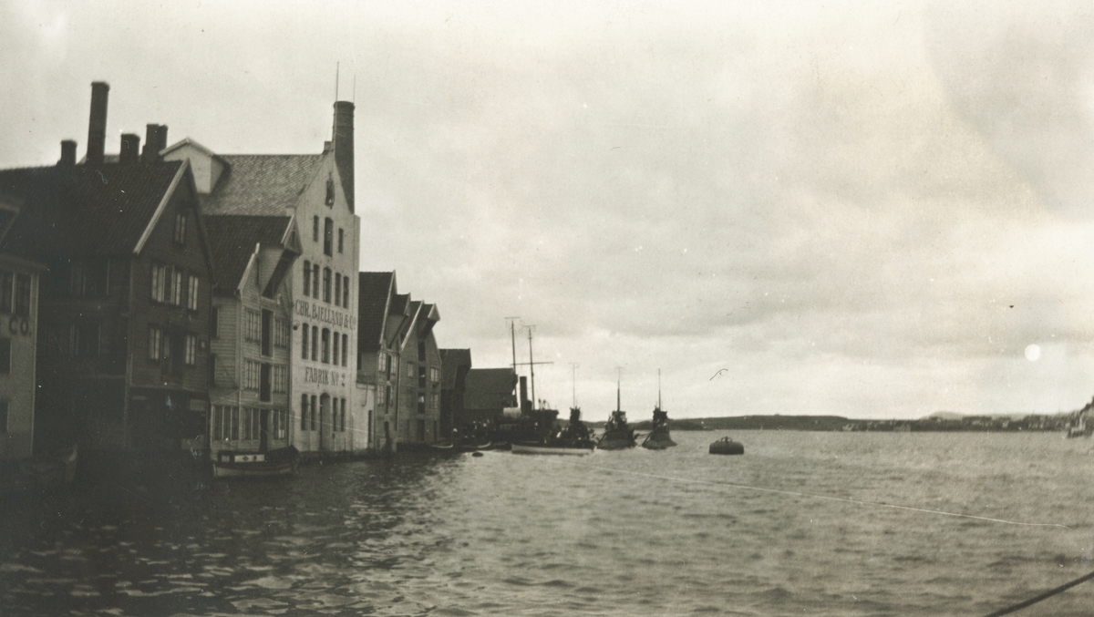 Motiv av havnemiljø ved ukjent sted. Flere båter ligger utenfor ei rekke med sjøboder. På ene bygningen står det "Chr. Bjelland & co. Fabrik nr. 2."