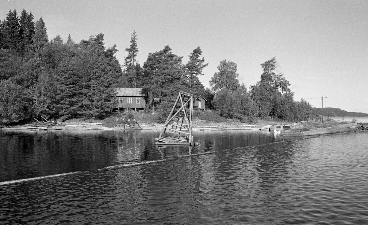 Fra Sleppetangen i Spydeberg i Østfold, ved den søndre delen av innsjøen Øyeren.  Fotografiet er tatt fra vannet mot land.  I et innlenset område mot stranda ligger en skjevtliggende flåte med et pyramidalt stillas.  Denne konstruksjonen skal ha fungert som lyskastertårn ytterst på ei trebrygge, forankret i ei steinfylt tømmerkiste.  Tårnet ble overflødig omkring 1960, da det ble støpt en ny molo hvor lyskasteren ble plassert. På stranda bakenfor skimter vi .  Litt høgere oppe på land skimter vi et saghus og skjul, omgitt av blandingsskog.  Til høyre i bildet ser vi et lite bryggeanlegg av betong.  Sleppetangen var oppankringssted for de store tømmerslepene som ble hentet fra Fetsund lenser i nordenden av innsjøen til Glommas utløp, der vaierne ble tatt av slepet.  Som regel gjaldt dette også «grimene» (vaierbindslene) rundt de enkelte «mosene» (tømmerbuntene) slepet besto av, slik at stokkene kunne flyte løst mot tømmerrennene forbi kraftverkene i vassdraget nedenfor.