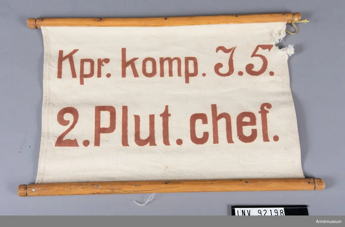 Skylt av tyg av smärtingtyp fästa i träpinnar på långsidorna. Röd text: "Kpr. komp. I.5. 2. Plut. chef."