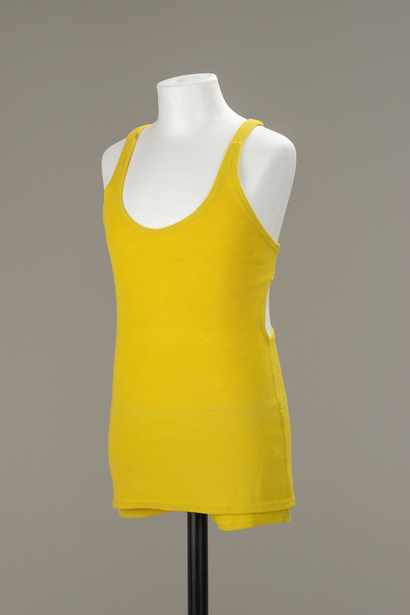 Sitrongul ullbadedrakt med shortsformede ben og over det et lite skjørt. Horisintalt bånd på tvers av ryggen og vertikal snor mellom skulderstropper og nedre del av badedrakten.