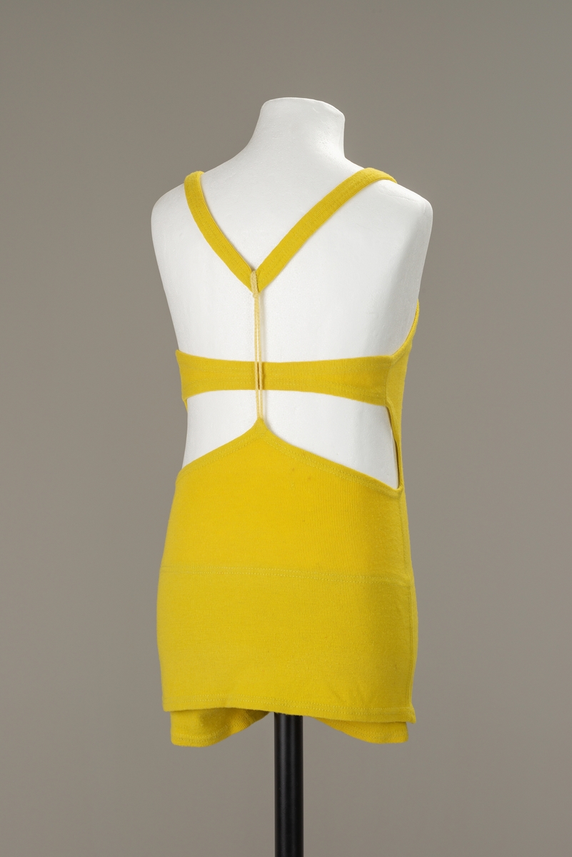 Sitrongul ullbadedrakt med shortsformede ben og over det et lite skjørt. Horisintalt bånd på tvers av ryggen og vertikal snor mellom skulderstropper og nedre del av badedrakten.