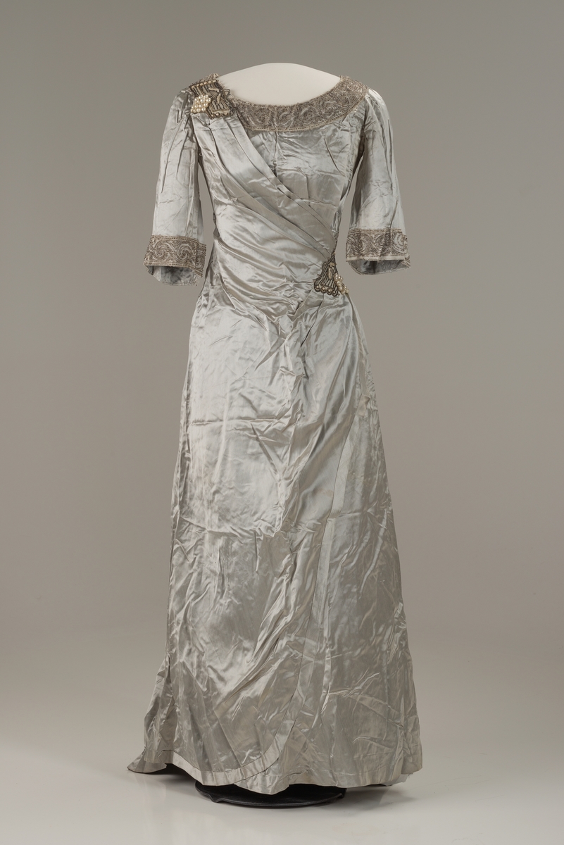 Skreddersydd, sølvfarget selskapskjole i kunstsilke med halvlange ermer og slep bak. Åpen, rund hals kantet med perlebroderi. Perlebroderi også nederst på ermene og til venstre i midjen. Flettet bån i kjolens stoff i ryggen. Øvre del av kjolen og ermene er fôret med bomull med isydde spiler av metall. Fôret lukkes med hekter. I fronten festes fôret i kjolen med trykknapper.