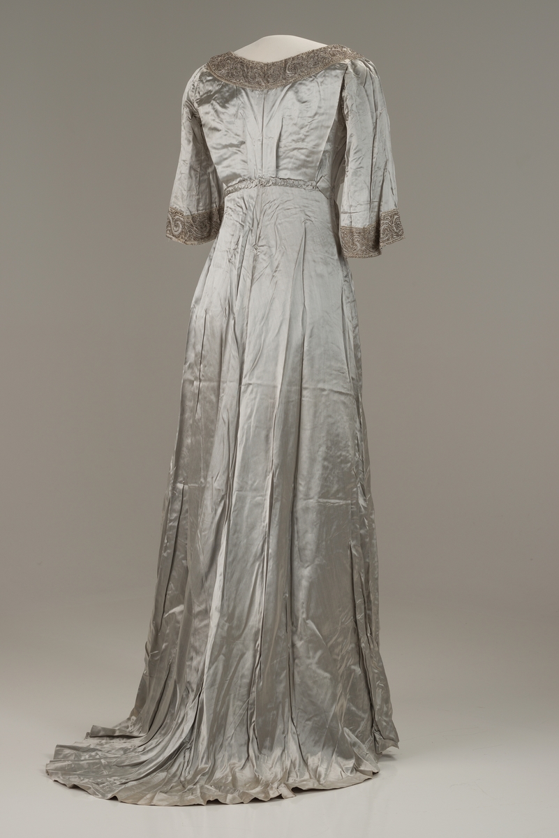 Skreddersydd, sølvfarget selskapskjole i kunstsilke med halvlange ermer og slep bak. Åpen, rund hals kantet med perlebroderi. Perlebroderi også nederst på ermene og til venstre i midjen. Flettet bån i kjolens stoff i ryggen. Øvre del av kjolen og ermene er fôret med bomull med isydde spiler av metall. Fôret lukkes med hekter. I fronten festes fôret i kjolen med trykknapper.