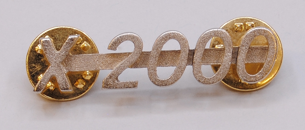 Silverfärgad pin. Denna pin är formad som texten "X 2000" och är gjuten med smal metallinje på baksidan som håller ihop texten. 

På baksidan av denna pin sitter två metallpiggar som ska träs igenom ett klädesplagg och fästas med fjärilslås.