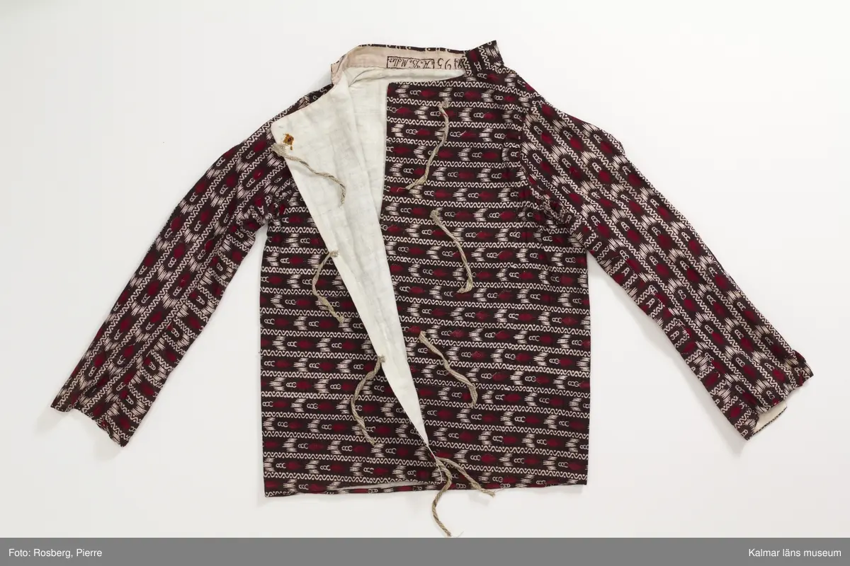 KLM 35481 Tröja. Sydd av kattuntryckt bomullstyg, svart botten med tvärgående ränder i bomullstygets bottenfärg samt i mörkrött. Tröjan är skuren i ett stycke utan sidsömmar. Lång ärm hopsydd av två delar med längsgående sömmar, den ena delen är skuren på längden den andra delen är skuren på tvären. Sprund vid handlederna, rund halsringning med ståkrage. Knyts framtill med gråvitrandiga band av linne. Fodrad med vitt bomullstyg. Halskantens foder av tygets avslutningskant med tryckt text: Blecktornet 1808 95 AB Möller. Tyget är tryckt på Blecktornets Kattuntryckeri på Södermalm, Stockholm.