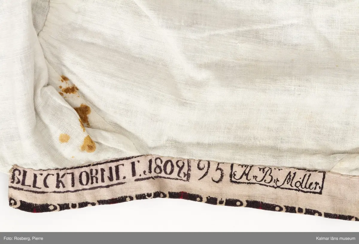 KLM 35481 Tröja. Sydd av kattuntryckt bomullstyg, svart botten med tvärgående ränder i bomullstygets bottenfärg samt i mörkrött. Tröjan är skuren i ett stycke utan sidsömmar. Lång ärm hopsydd av två delar med längsgående sömmar, den ena delen är skuren på längden den andra delen är skuren på tvären. Sprund vid handlederna, rund halsringning med ståkrage. Knyts framtill med gråvitrandiga band av linne. Fodrad med vitt bomullstyg. Halskantens foder av tygets avslutningskant med tryckt text: Blecktornet 1808 95 AB Möller. Tyget är tryckt på Blecktornets Kattuntryckeri på Södermalm, Stockholm.