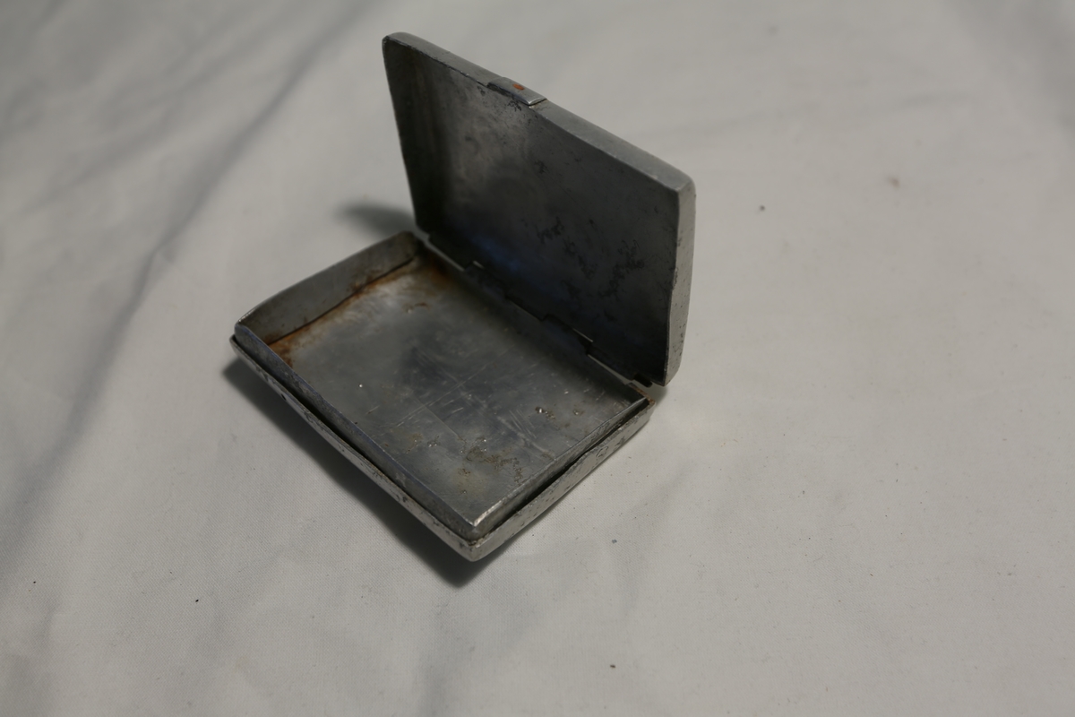 Kjærestepar i rombisk ramme med ornamenter samt bokstavene K og S (speilvendt) på lokket. Kystlandskap med båt og lav sol over fjell i oval ramme med snirklede ornamenter.